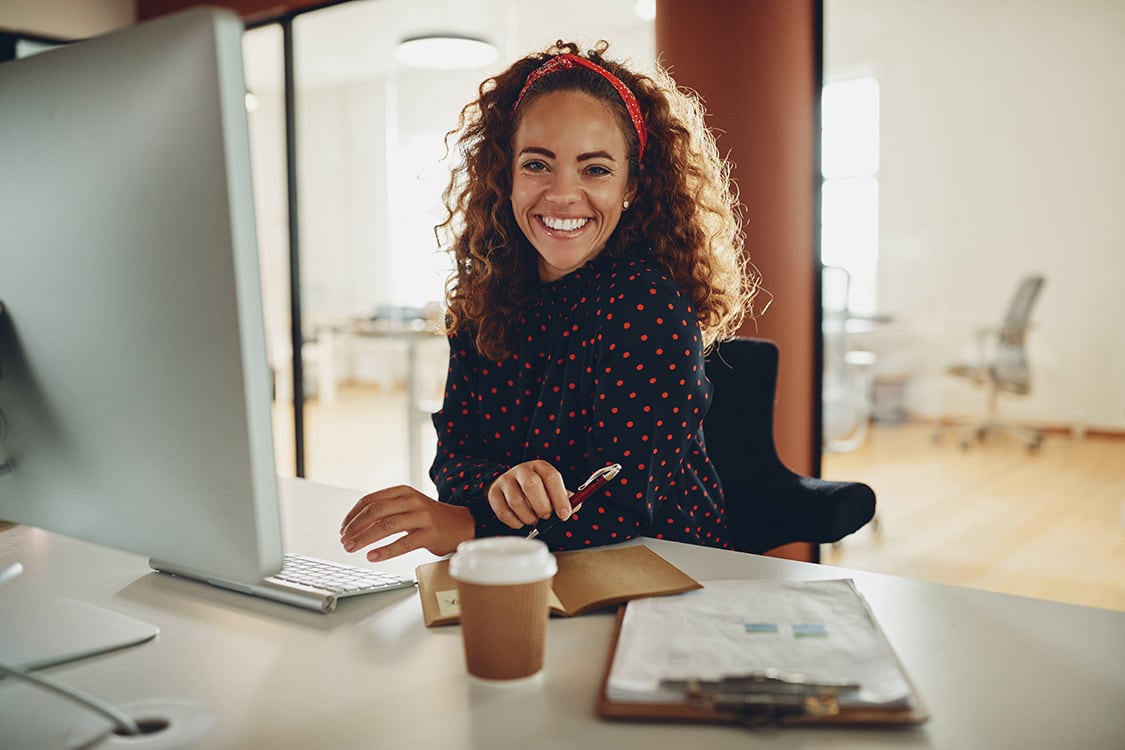 A smiling woman with curly red hair at a desk with a computer, takeaway coffee cup and clipboard in a bright open-plan office