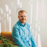 A smiling man in a light blue hoodie seated in front of a white wall
