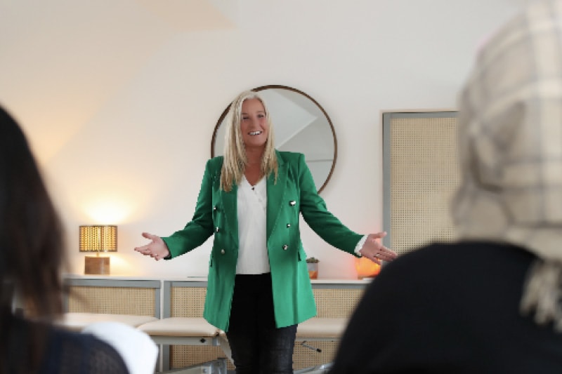 A woman in a green blazer speaking to a small seated audience in a bright, modern interior room