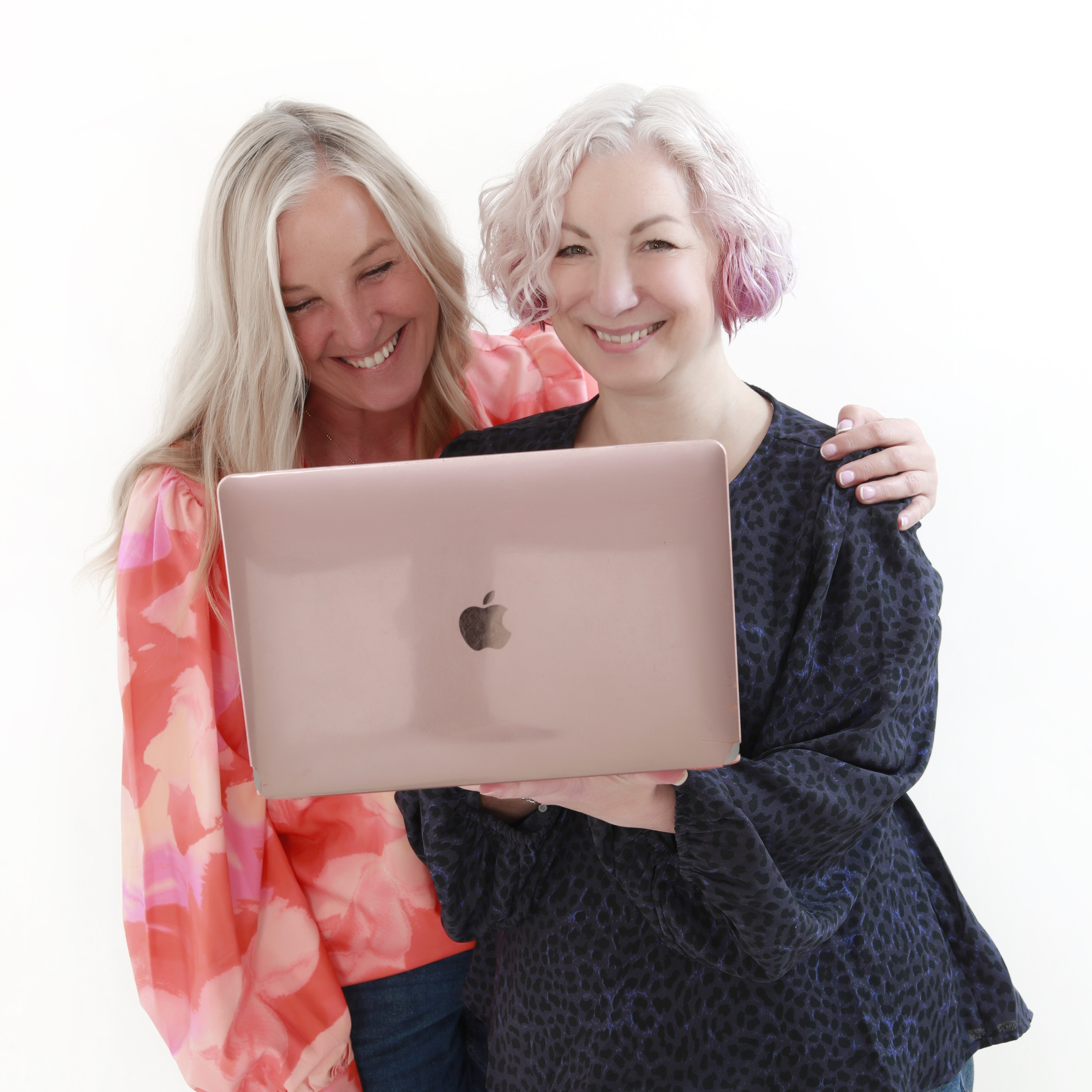 Michelle and Rachael smiling together, with Rachael holding a rose-gold MacBook, against a plain white studio background.