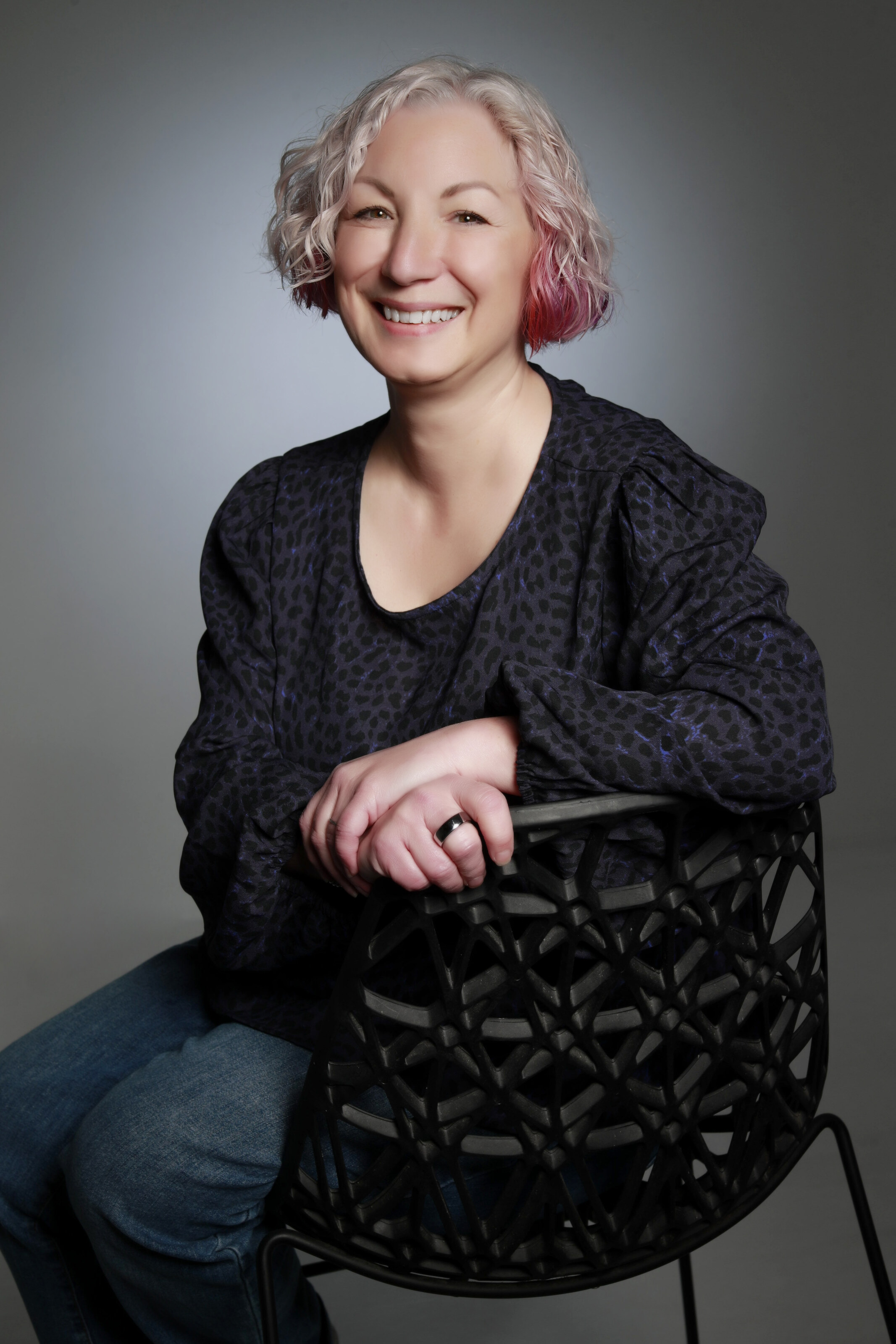 Rachael, smiling, with short curly silver and pink hair, seated astride a black lattice chair against a grey studio background.