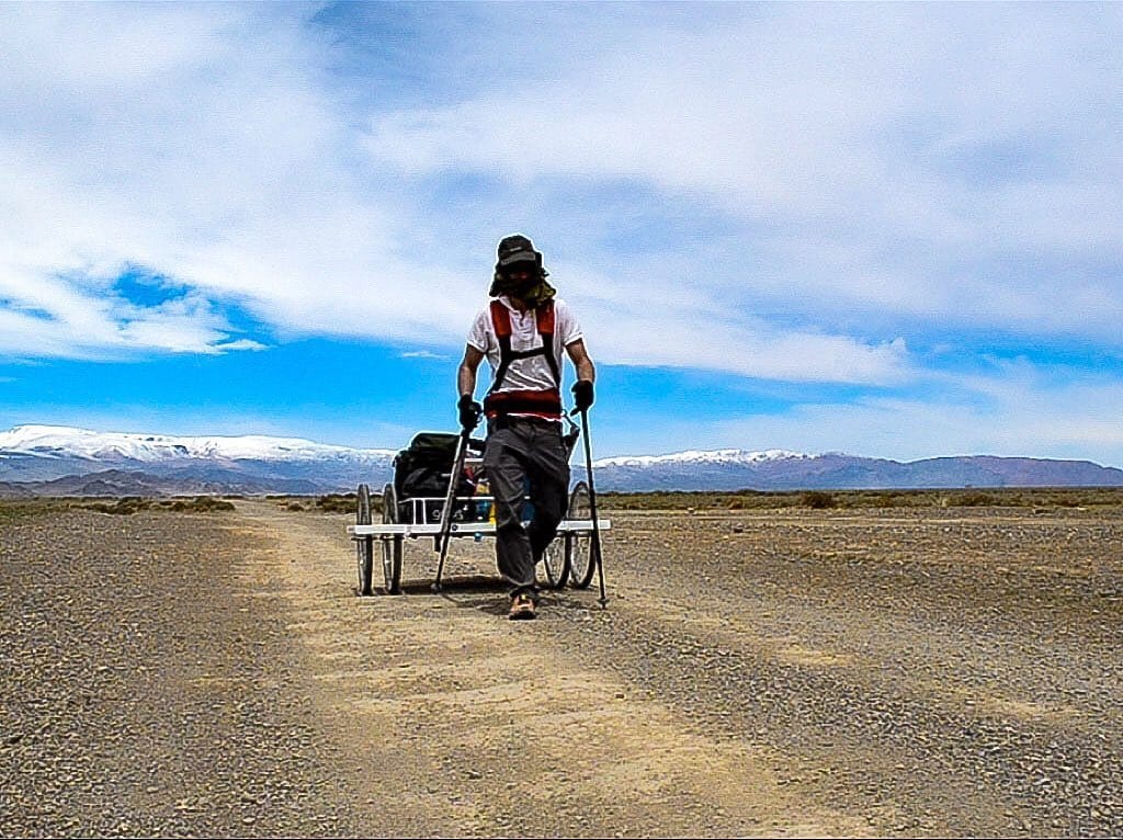 Ash Dykes hauling a 120kg wheeled trailer across the Gobi Desert, snow-capped Altai Mountains behind him