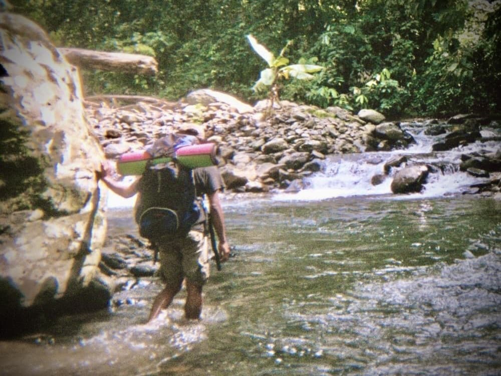 a hiker wading through a shallow jungle river with a fully loaded backpack
