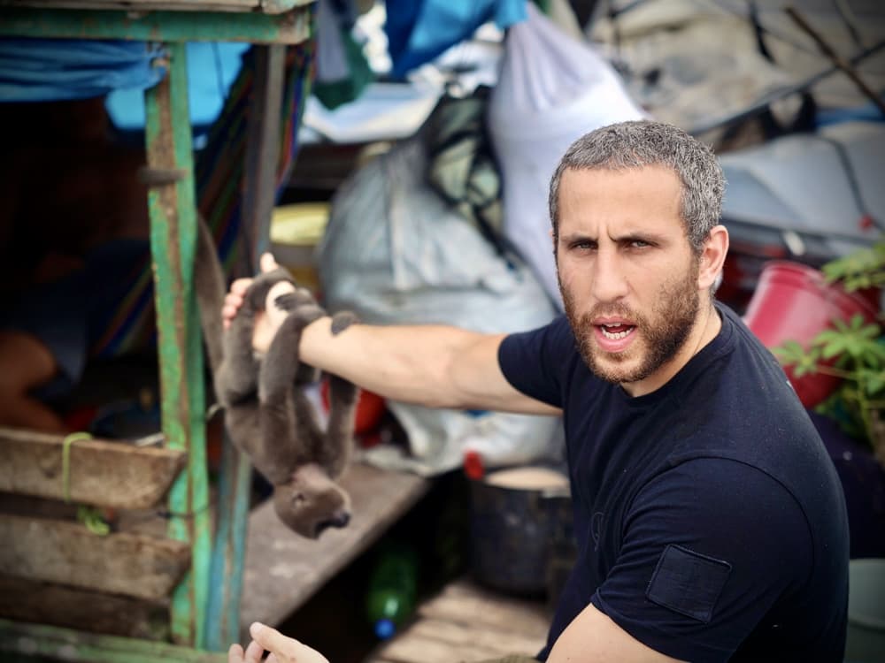 a man holding a small wild animal, surrounded by cluttered equipment in an outdoor setting