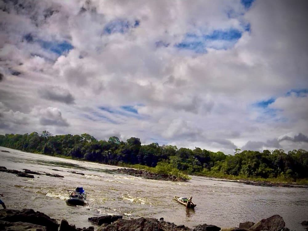 two loaded motorboats on a wide jungle river with rapids and rocky shoreline
