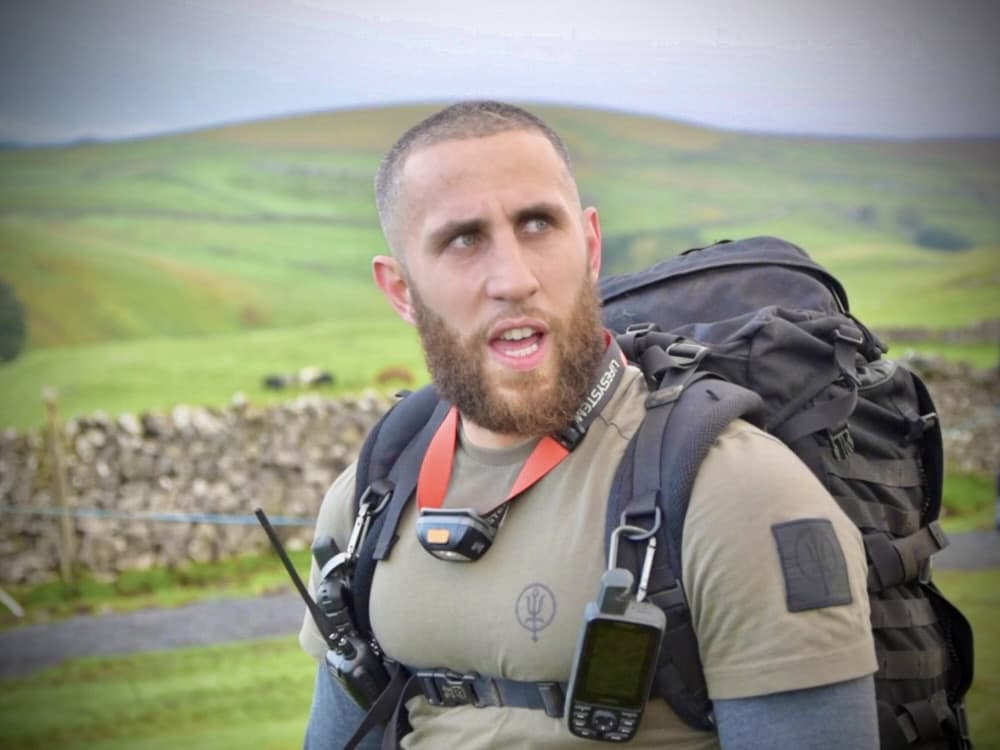 close-up portrait of a man in tactical gear with a large rucksack, GPS unit, and radio, outdoors against rolling green hills and a dry stone wall