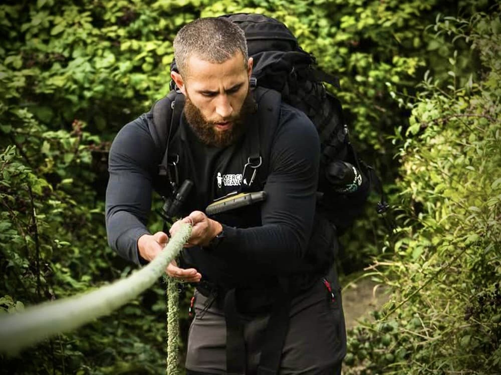 a man with a heavy backpack gripping a moss-covered guide rope on a wooded trail