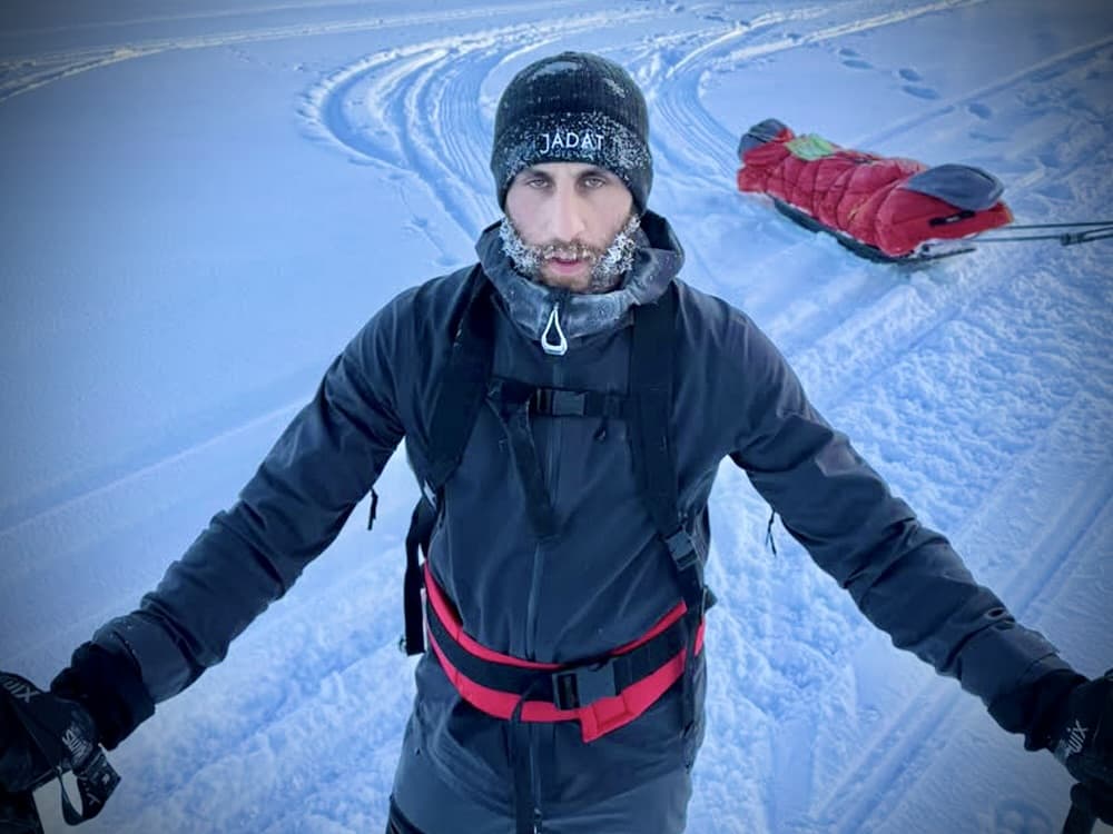 a man in expedition cold-weather gear pulling a loaded red pulk sled across snow, frost visible on his beard, holding ski poles