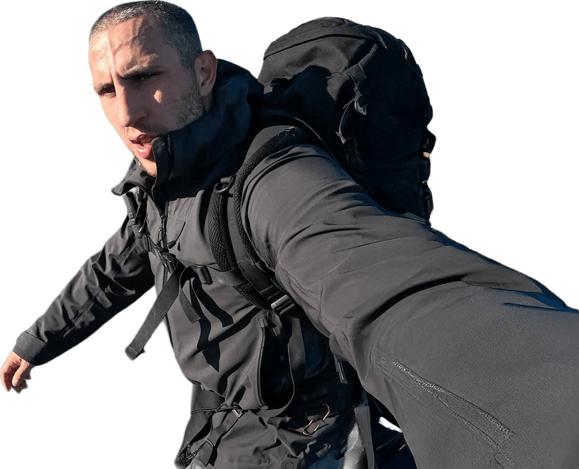 close-up selfie of a man carrying a large black hiking backpack, shot from a low angle on a white background