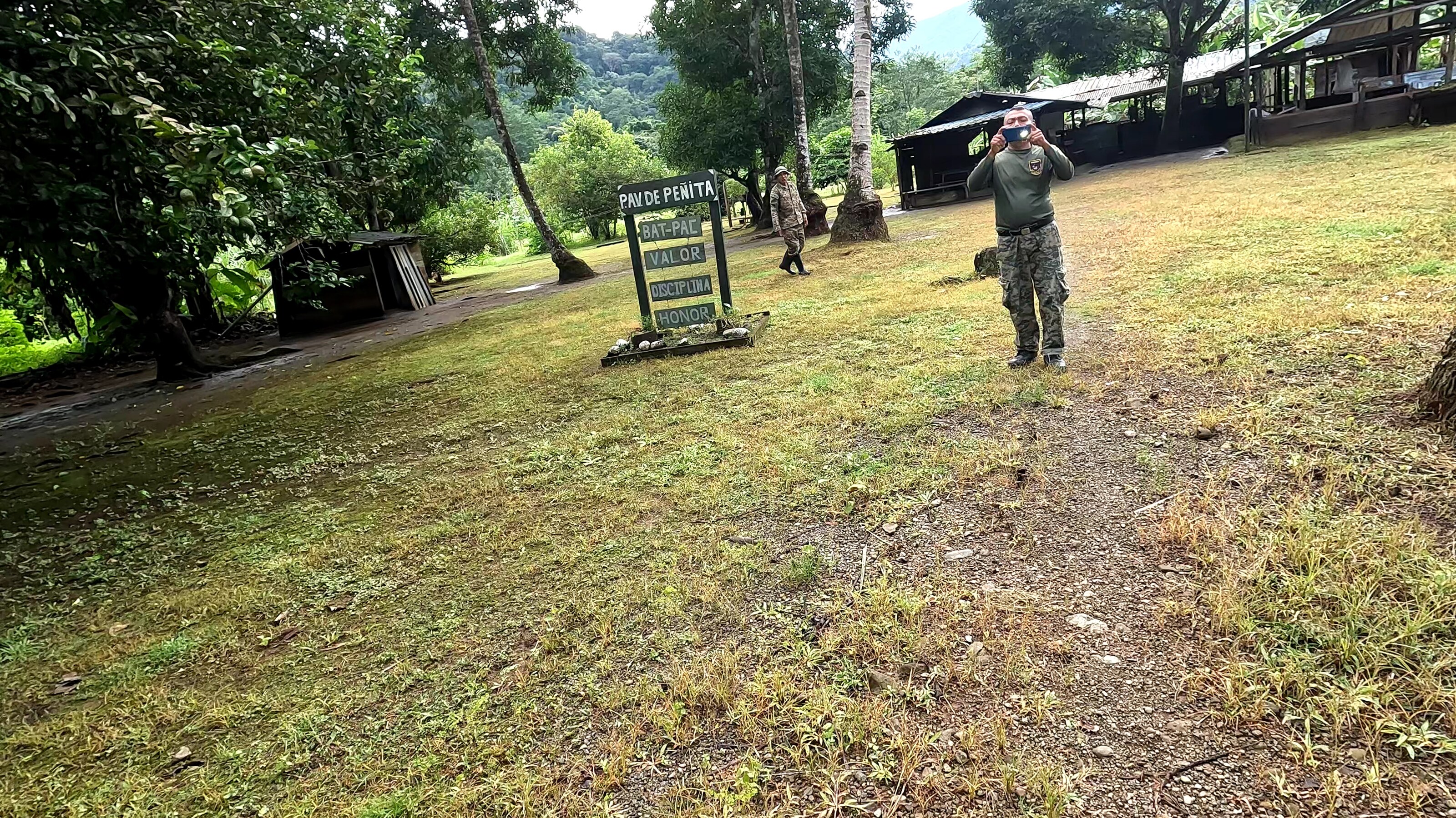 two uniformed personnel at an outdoor compound sign reading "PAV DE PENITA" with words valor, disciplina, and honor, surrounded by trees and open grass