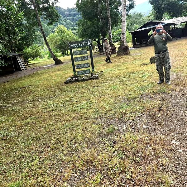 two uniformed personnel at an outdoor compound sign reading "PAV DE PENITA" with words valor, disciplina, and honor, surrounded by trees and open grass