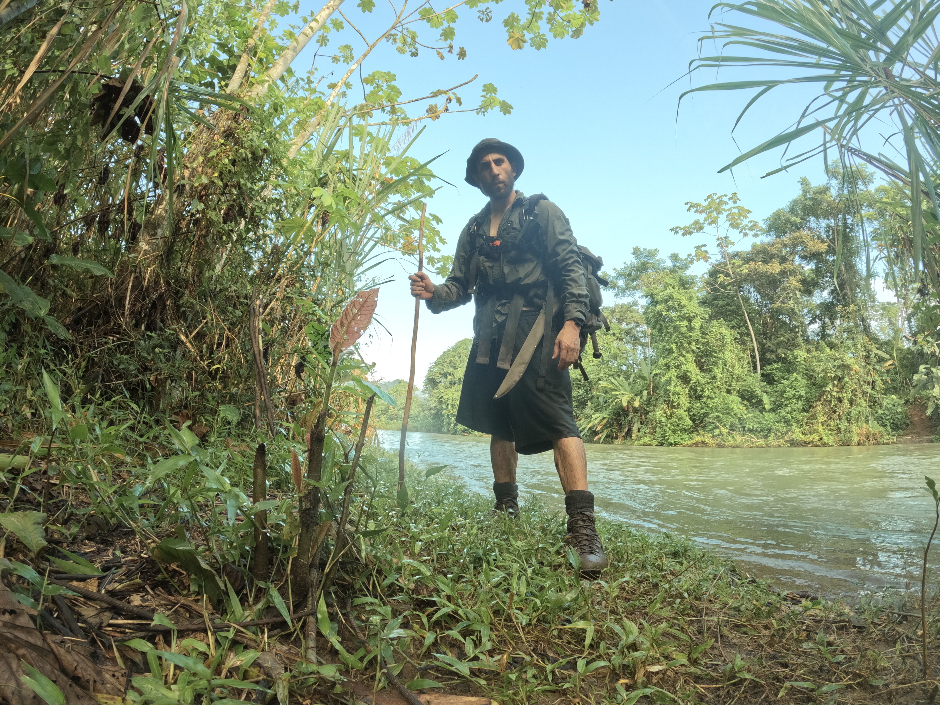 a man with a backpack, machete and walking stick standing on a dense jungle riverbank