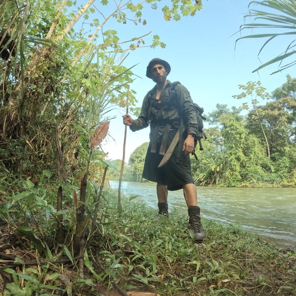 a man with a backpack, machete and walking stick standing on a dense jungle riverbank