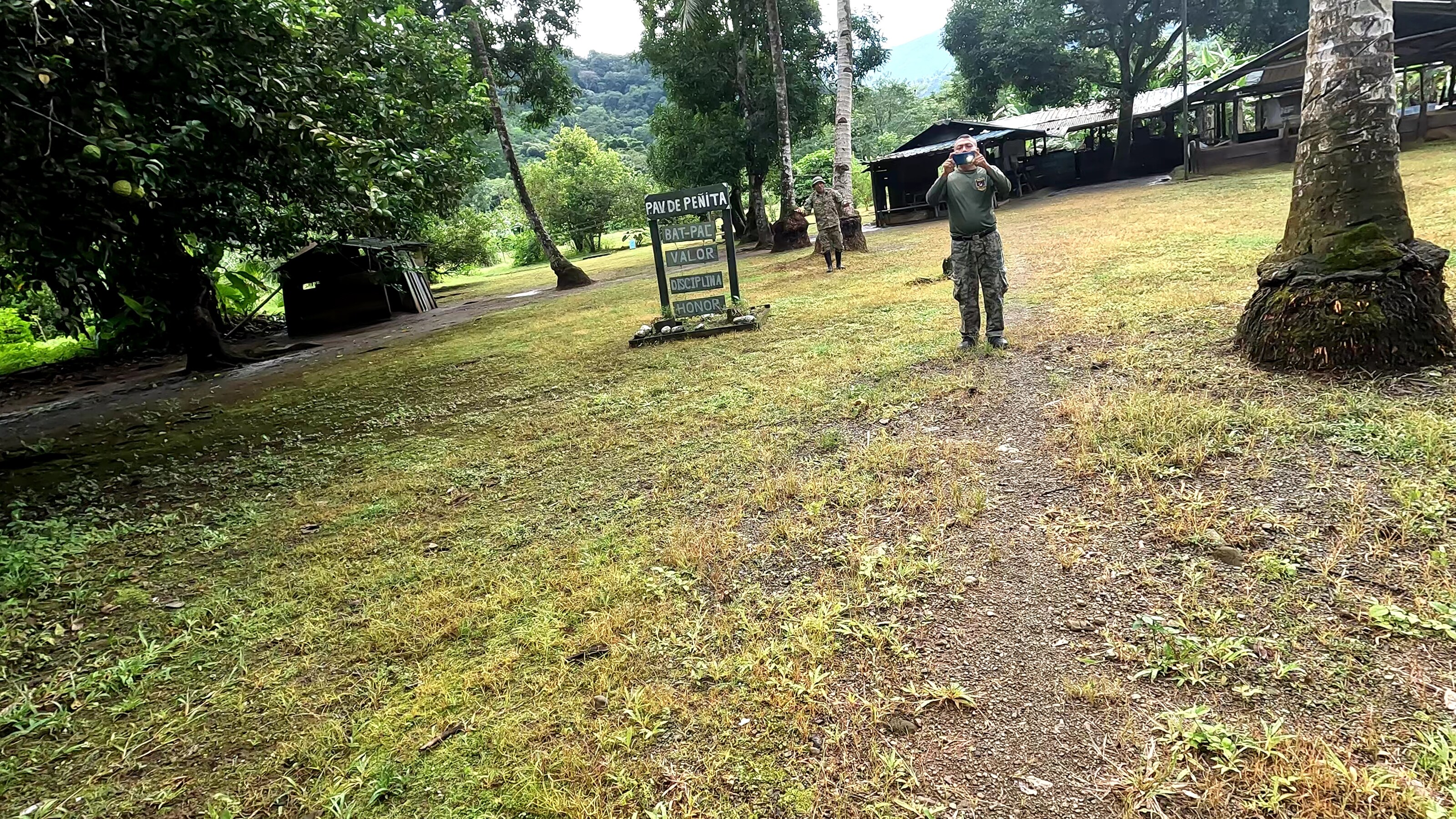 two uniformed personnel on a grassy outdoor compound with a military-style sign and jungle vegetation in the background