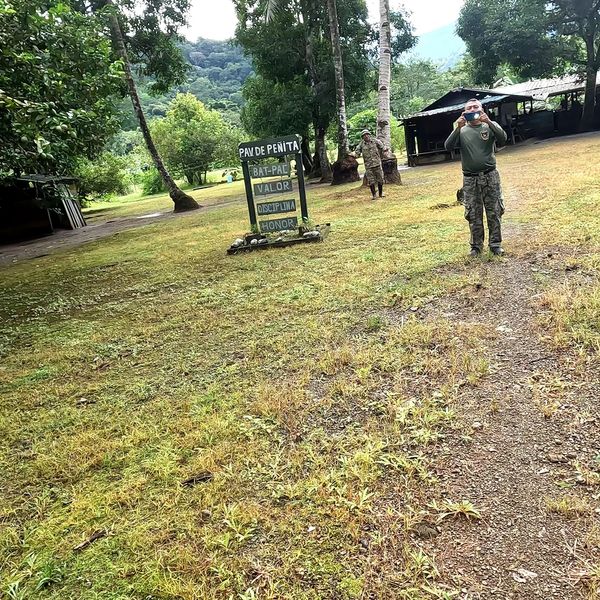 two uniformed personnel on a grassy outdoor compound with a military-style sign and jungle vegetation in the background