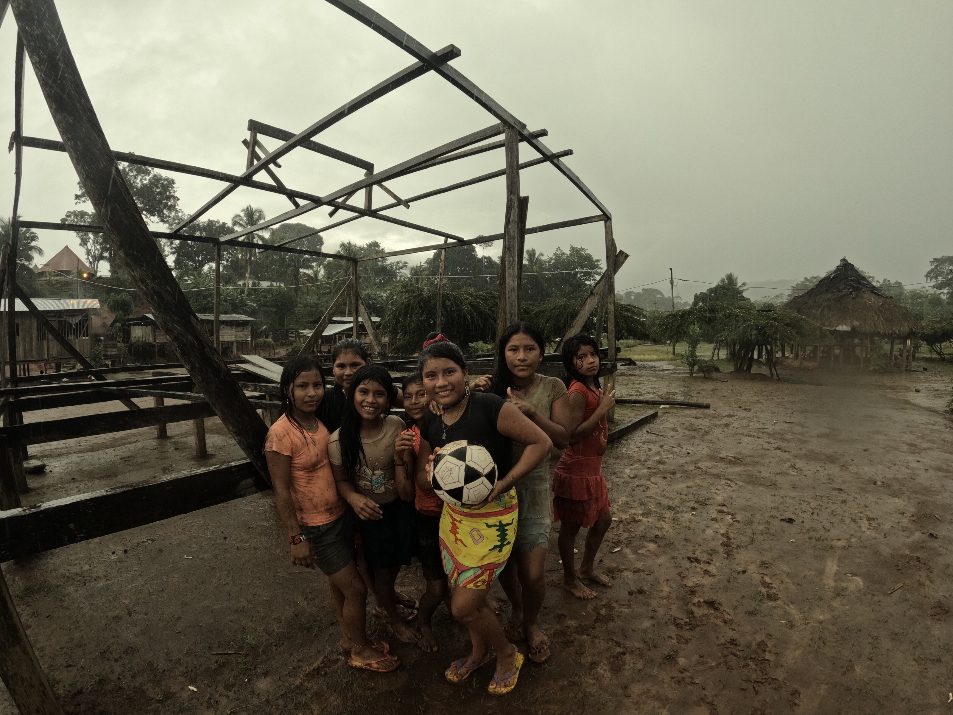 a group of children posing outdoors in heavy rain, holding a football, with an open wooden frame structure and thatched-roof buildings in the background