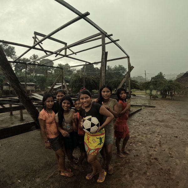 a group of children posing outdoors in heavy rain, holding a football, with an open wooden frame structure and thatched-roof buildings in the background