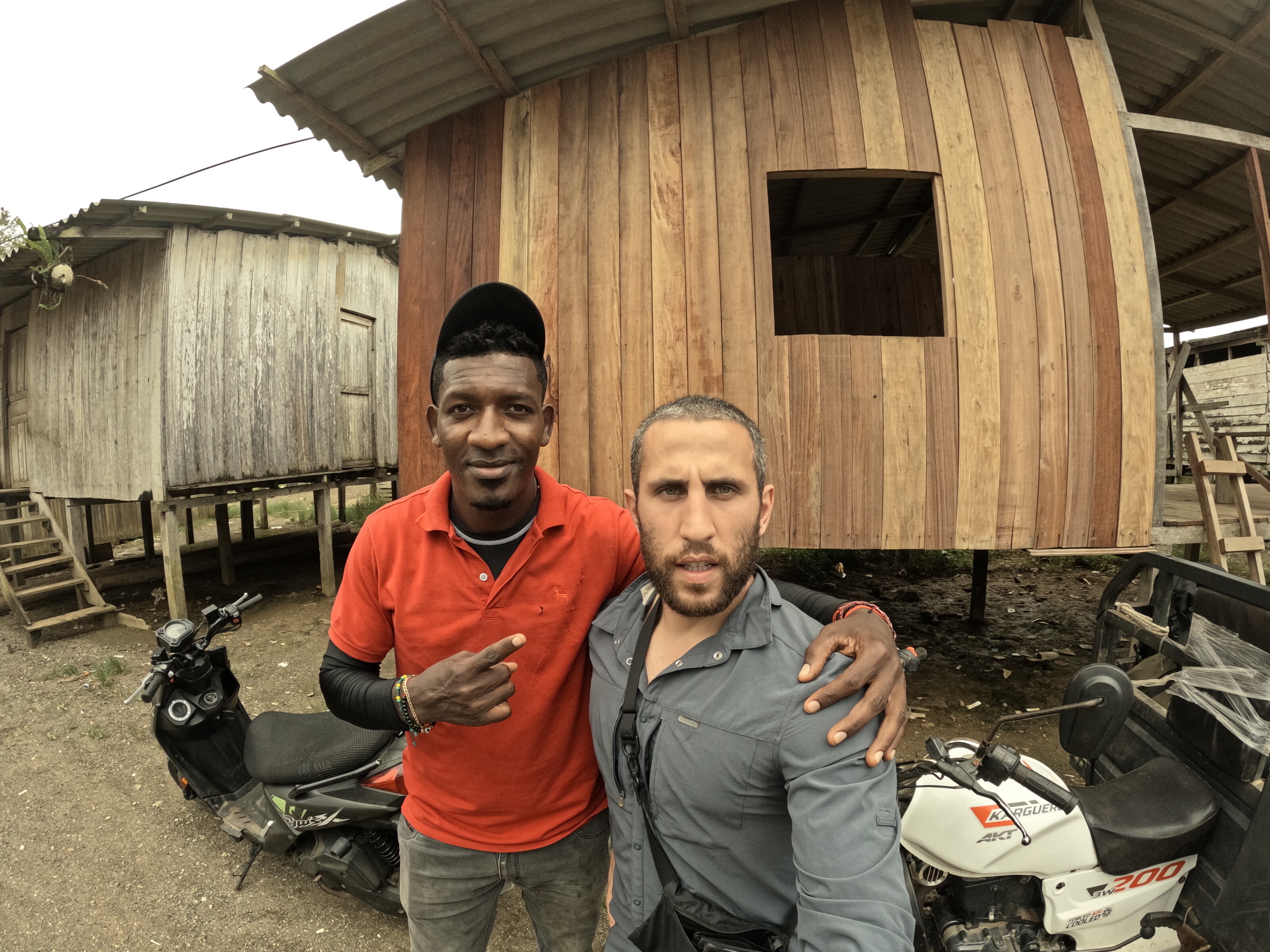 two men posing together outdoors beside motorcycles and wooden structures