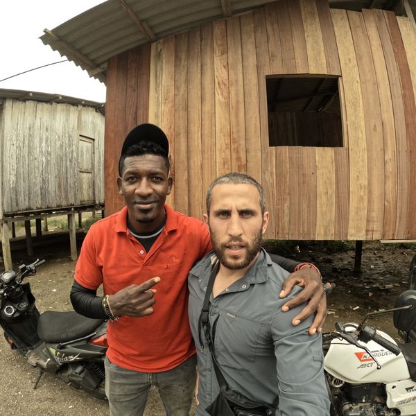 two men posing together outdoors beside motorcycles and wooden structures