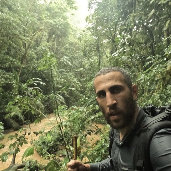 a man with a backpack taking a selfie in heavy rain inside a dense jungle, with a swollen muddy river visible behind him