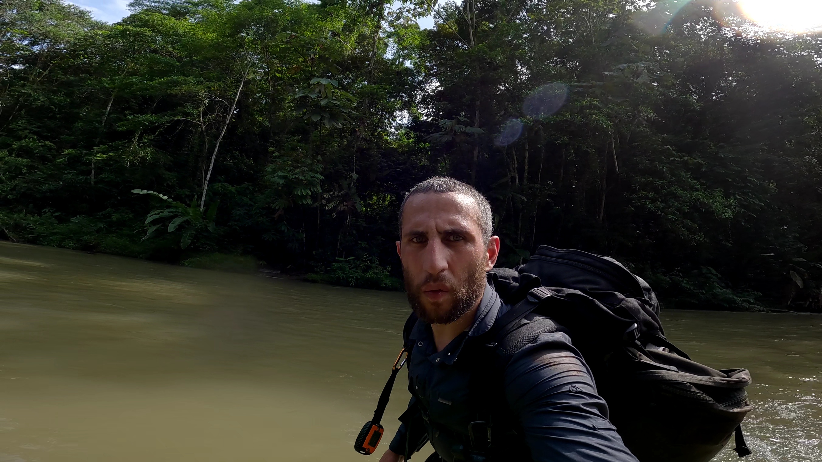 a bearded man with a large black backpack taking a selfie while standing in a murky jungle river, dense tropical forest in the background