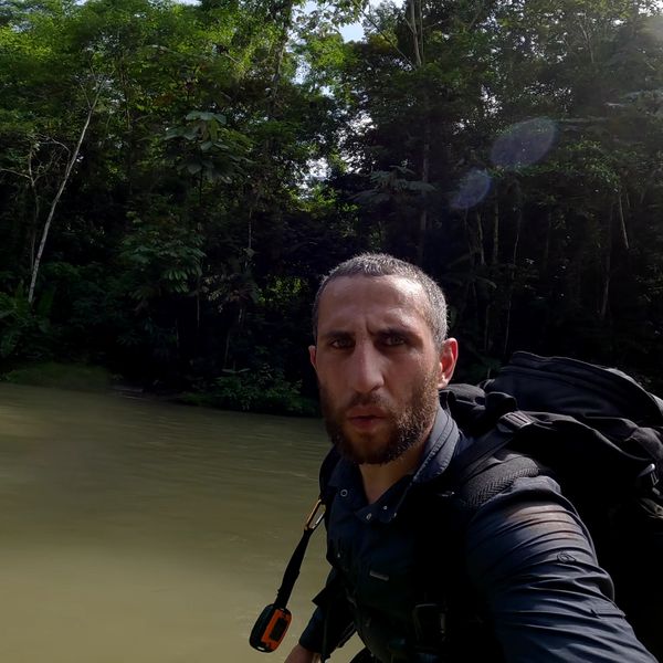 a bearded man with a large black backpack taking a selfie while standing in a murky jungle river, dense tropical forest in the background