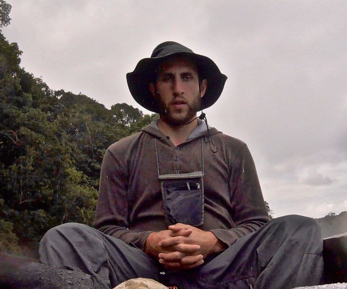 a man in outdoor gear sitting cross-legged on rocky ground in an overcast natural setting