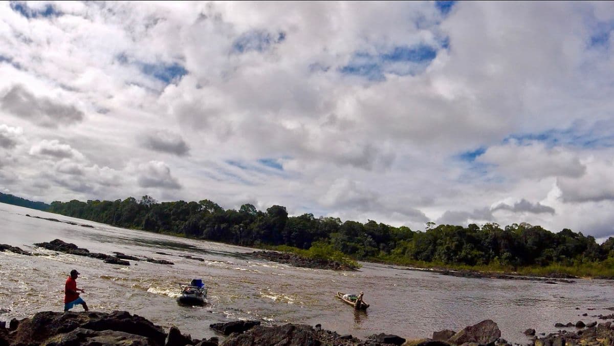 two small boats being guided through river rapids with a person on the rocky bank, dense forest treeline and cloudy sky in the background - wide landscape shot