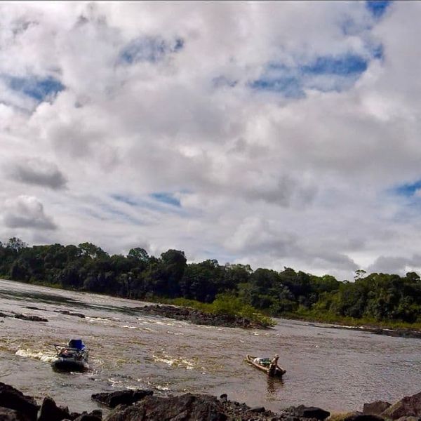 two small boats being guided through river rapids with a person on the rocky bank, dense forest treeline and cloudy sky in the background - wide landscape shot