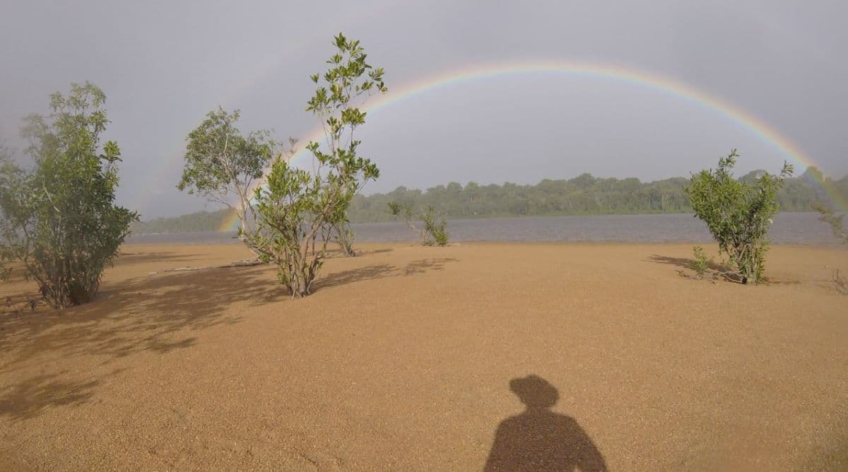 a double rainbow over a sandy tidal flat with low shrubs and a treeline in the distance, photographer's shadow visible in the foreground