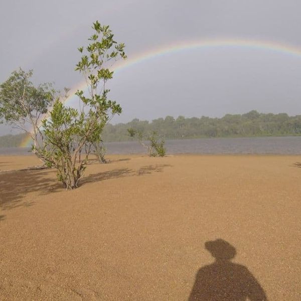a double rainbow over a sandy tidal flat with low shrubs and a treeline in the distance, photographer's shadow visible in the foreground