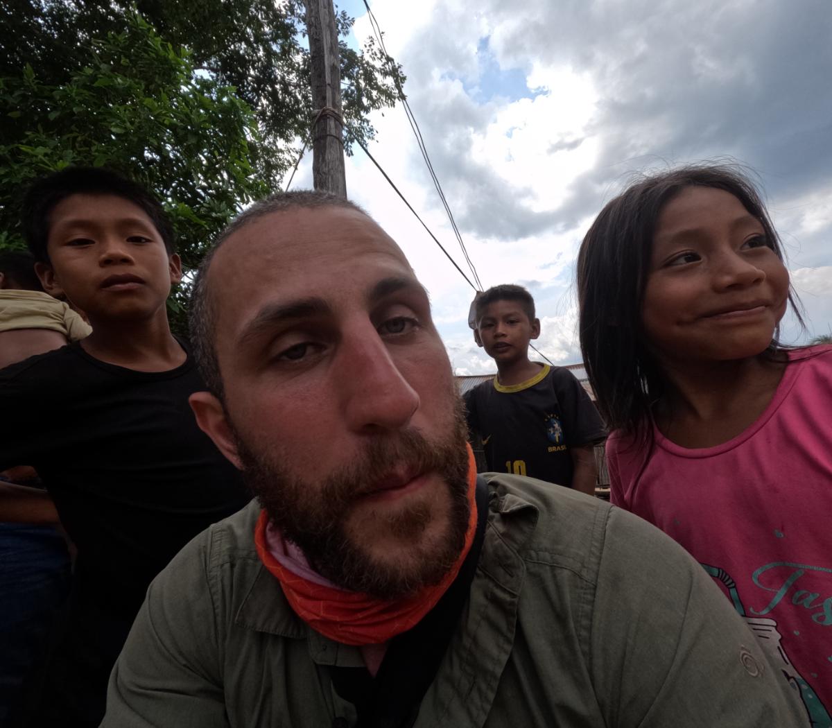 a bearded man taking a selfie outdoors with several children around him, cloudy sky and trees in the background