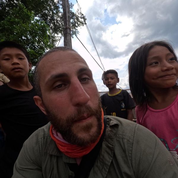 a bearded man taking a selfie outdoors with several children around him, cloudy sky and trees in the background