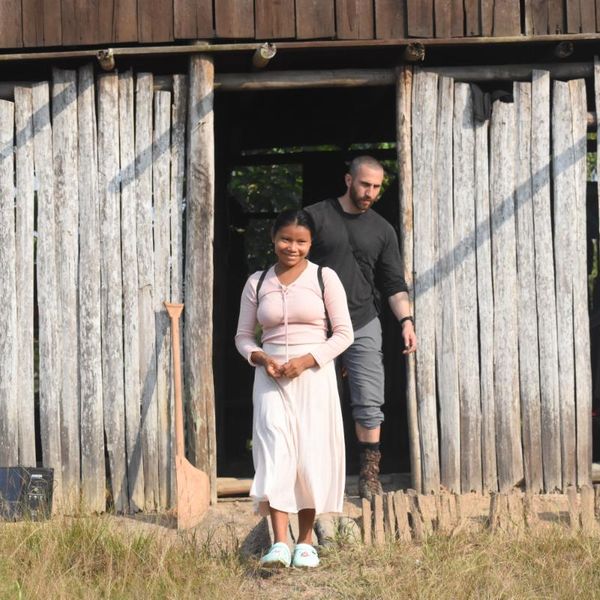 two people emerging from the doorway of a weathered timber building, with a wooden shovel leaning against the wall beside them