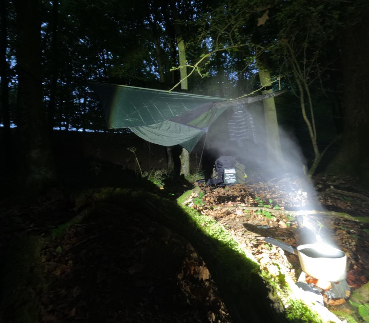 nighttime hammock and tarp camp setup in woodland with cooking pot and backpack