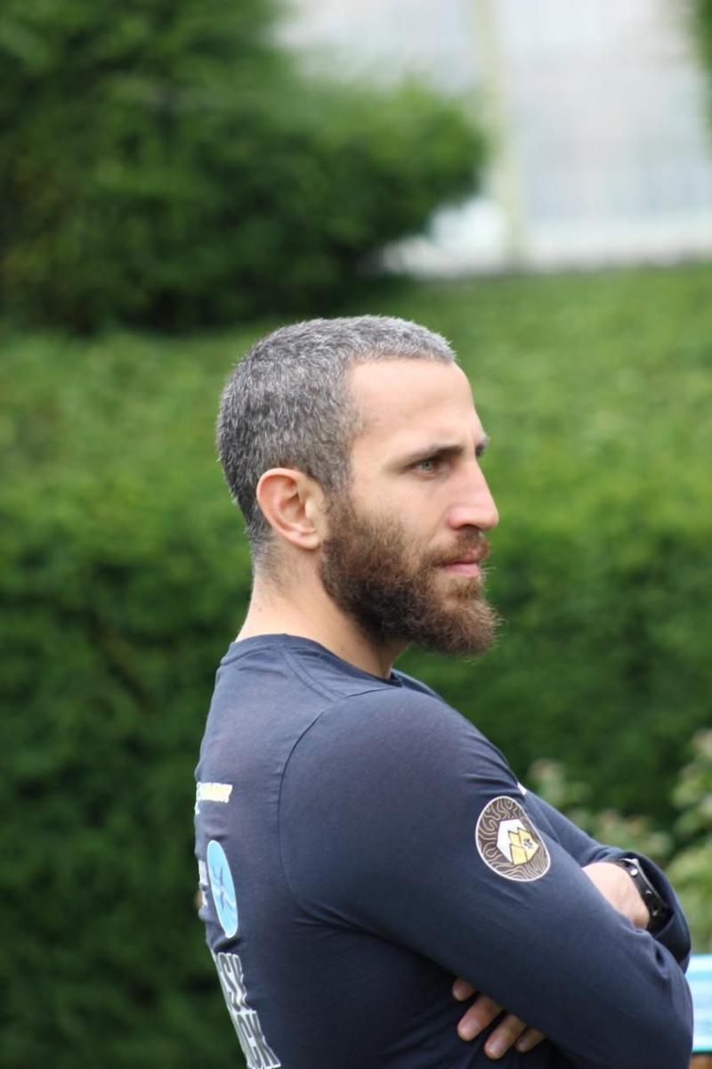 profile portrait of a man with a beard and crossed arms wearing a branded long-sleeve top, outdoors against a blurred green hedge background