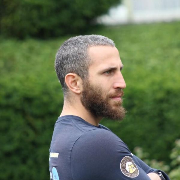 profile portrait of a man with a beard and crossed arms wearing a branded long-sleeve top, outdoors against a blurred green hedge background
