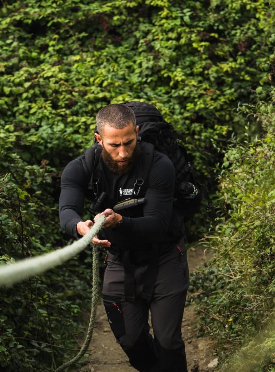 a man hauling himself up a steep forested trail using a fixed guide rope, wearing a large black backpack and all-black technical clothing
