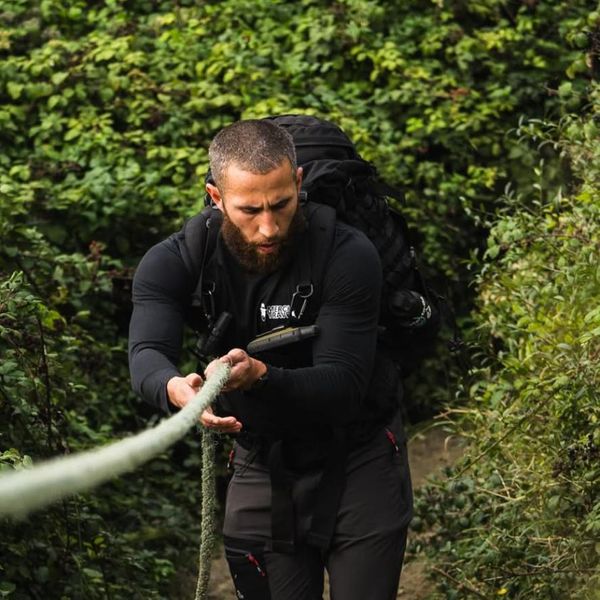 a man hauling himself up a steep forested trail using a fixed guide rope, wearing a large black backpack and all-black technical clothing