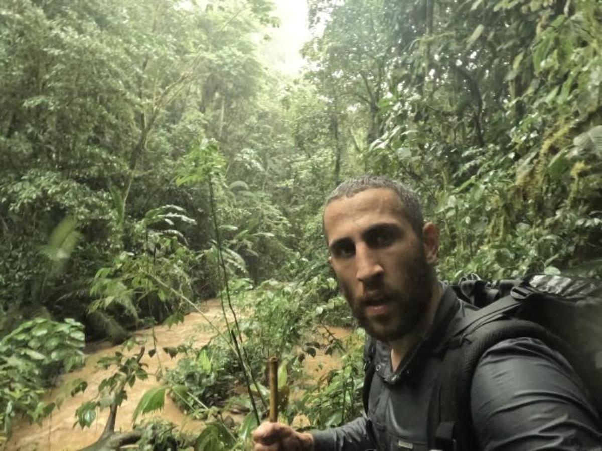 a man with a backpack and walking stick taking a selfie in dense jungle beside a swollen muddy river