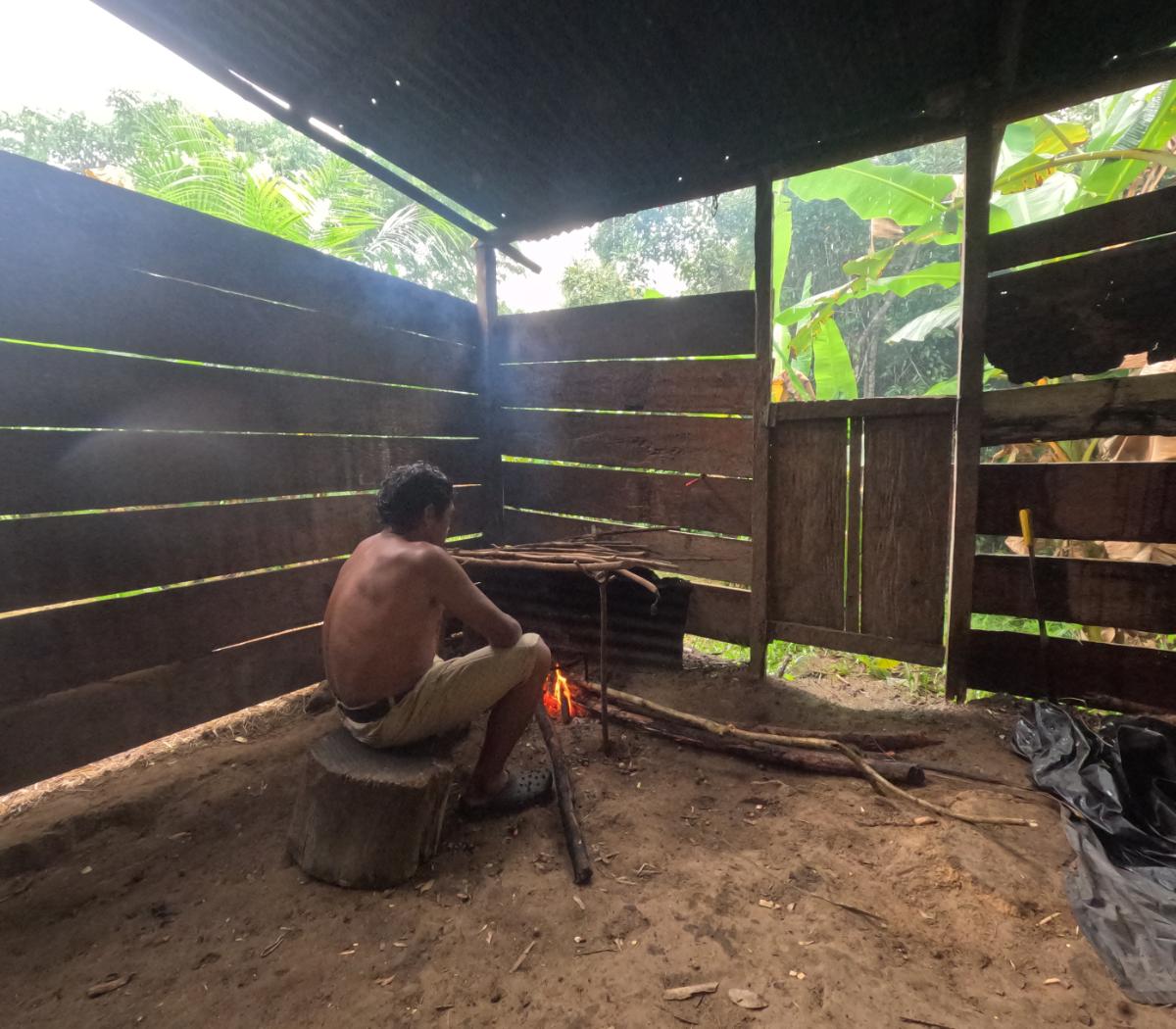 a man tending an open fire inside a traditional open-sided wooden shelter with tropical forest visible through the slatted walls
