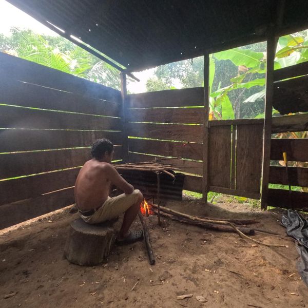 a man tending an open fire inside a traditional open-sided wooden shelter with tropical forest visible through the slatted walls