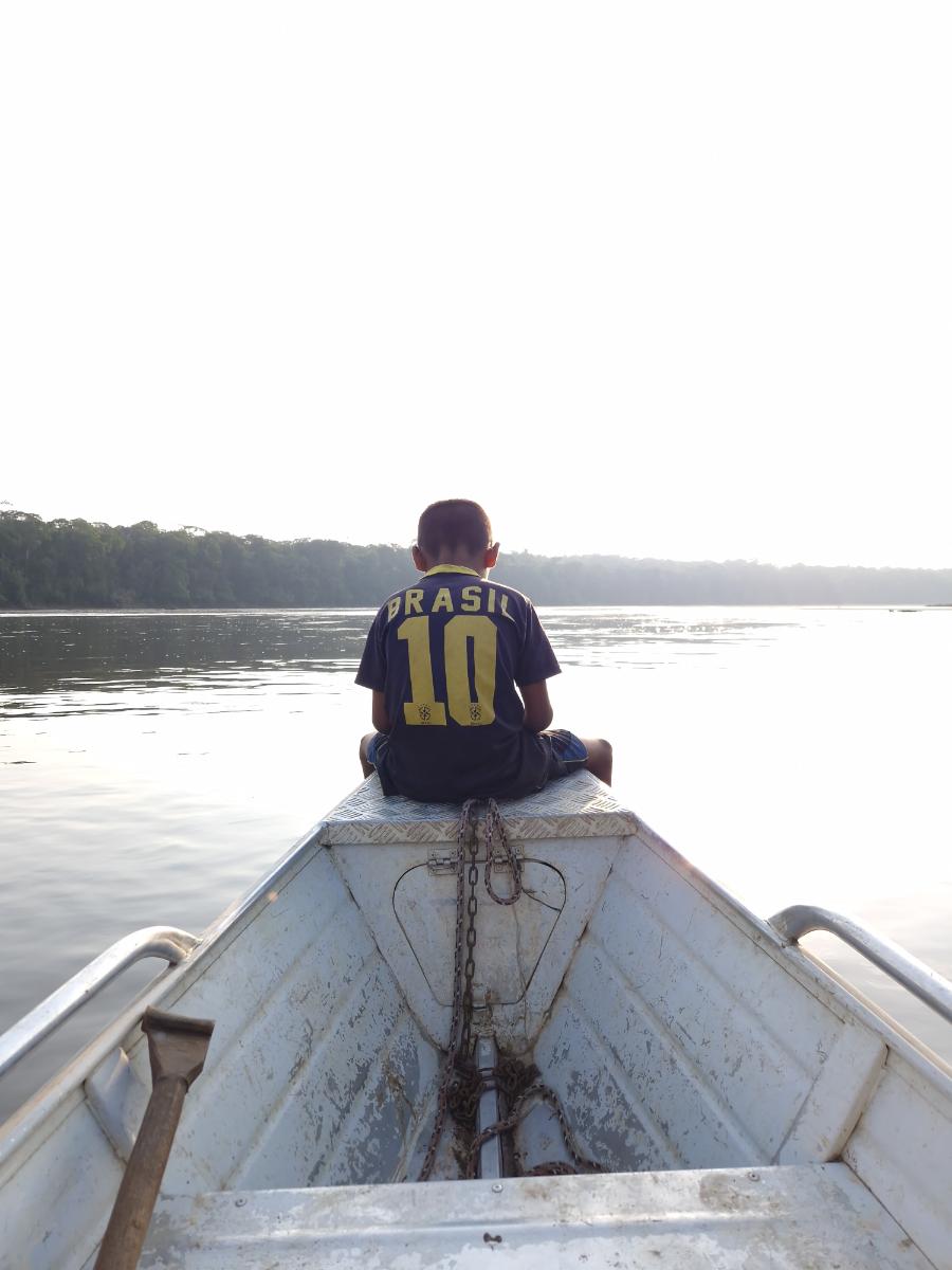 a child seated on the bow of a small aluminium boat looking out over a wide river, dense treeline in the hazy distance