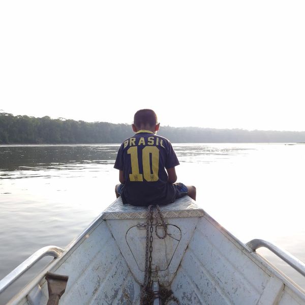 a child seated on the bow of a small aluminium boat looking out over a wide river, dense treeline in the hazy distance