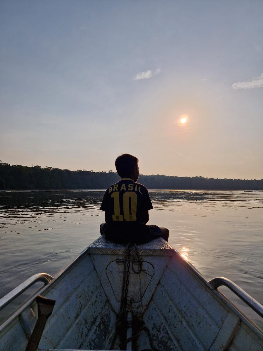 a child seated on a boat bow looking out over a calm river at sunset, dense forest treeline in the background