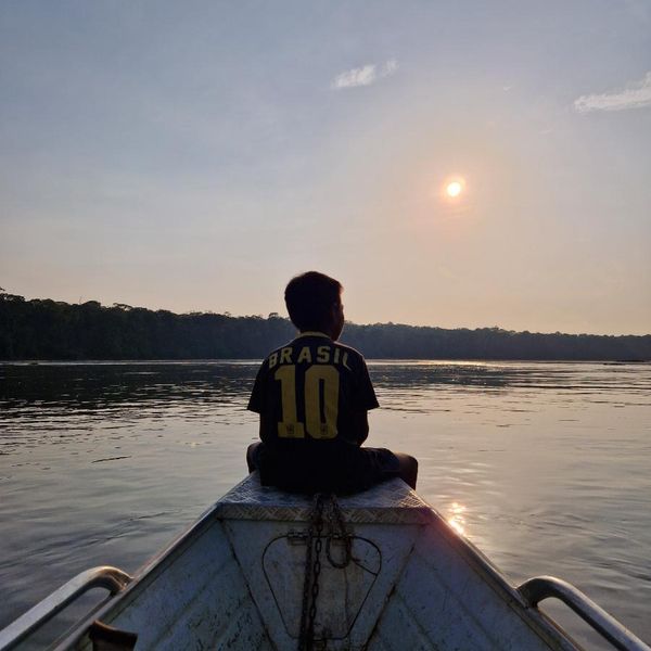 a child seated on a boat bow looking out over a calm river at sunset, dense forest treeline in the background