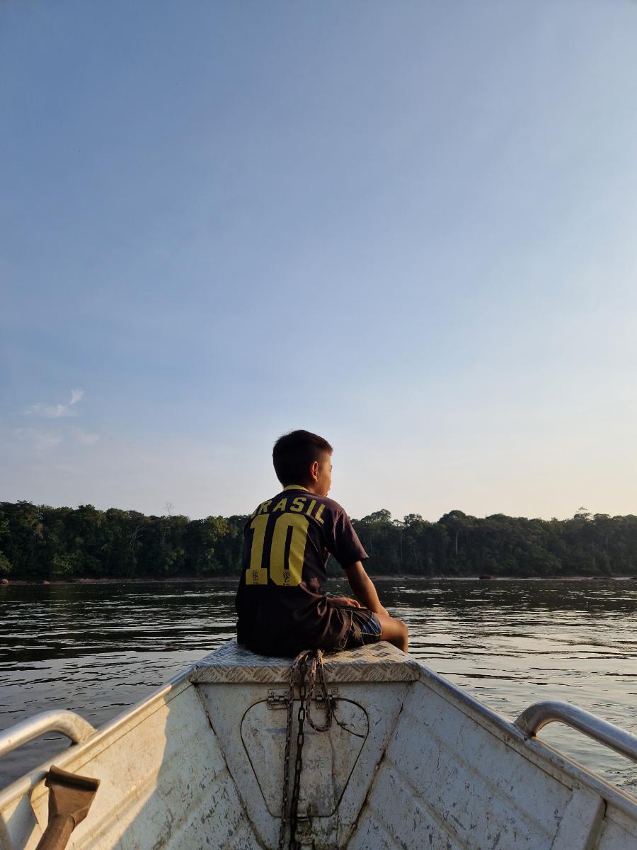 a child seated on the bow of a small motorboat gazing across a wide river with a forested far bank under a clear sky