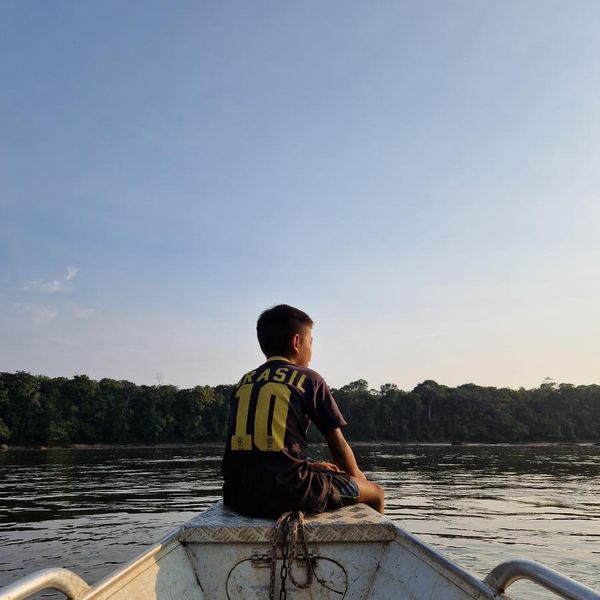 a child seated on the bow of a small motorboat gazing across a wide river with a forested far bank under a clear sky