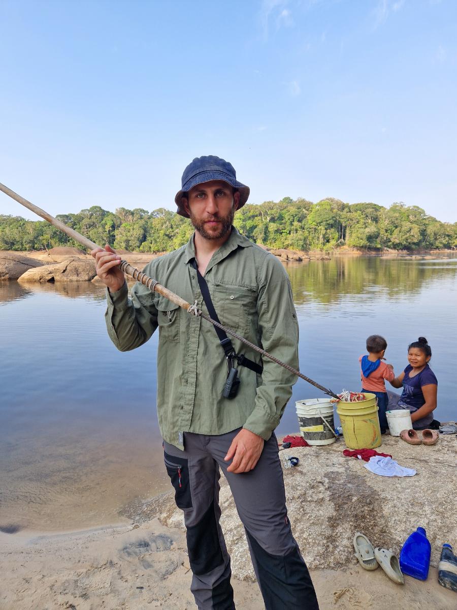 a man holding a traditional wooden spear on a rocky riverbank, with dense forest and two other people visible behind him