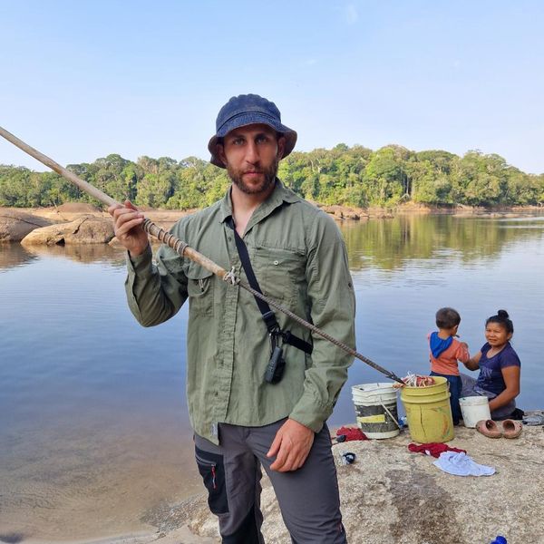 a man holding a traditional wooden spear on a rocky riverbank, with dense forest and two other people visible behind him